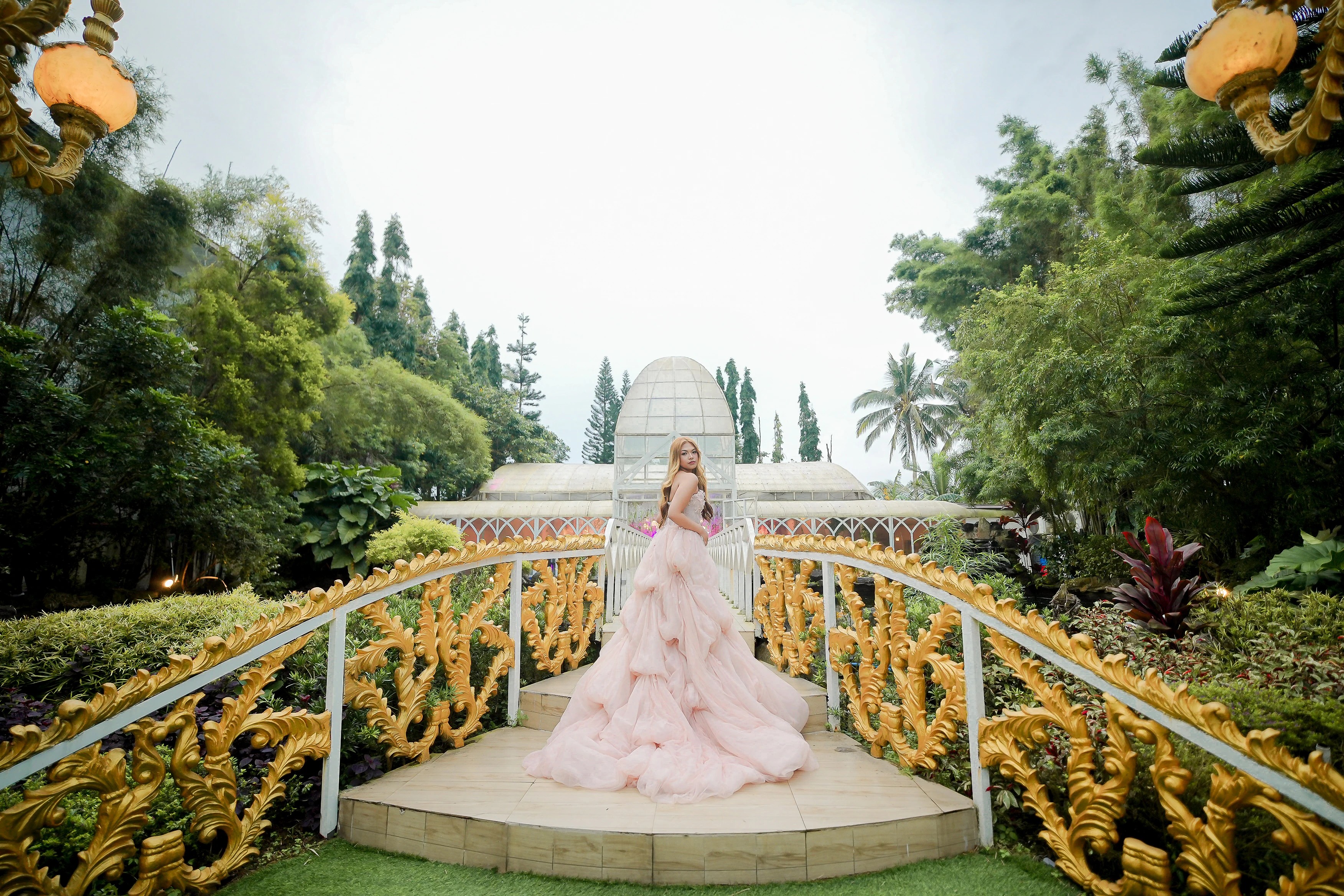 A woman in a flowing pink gown on a garden bridge surrounded by trees