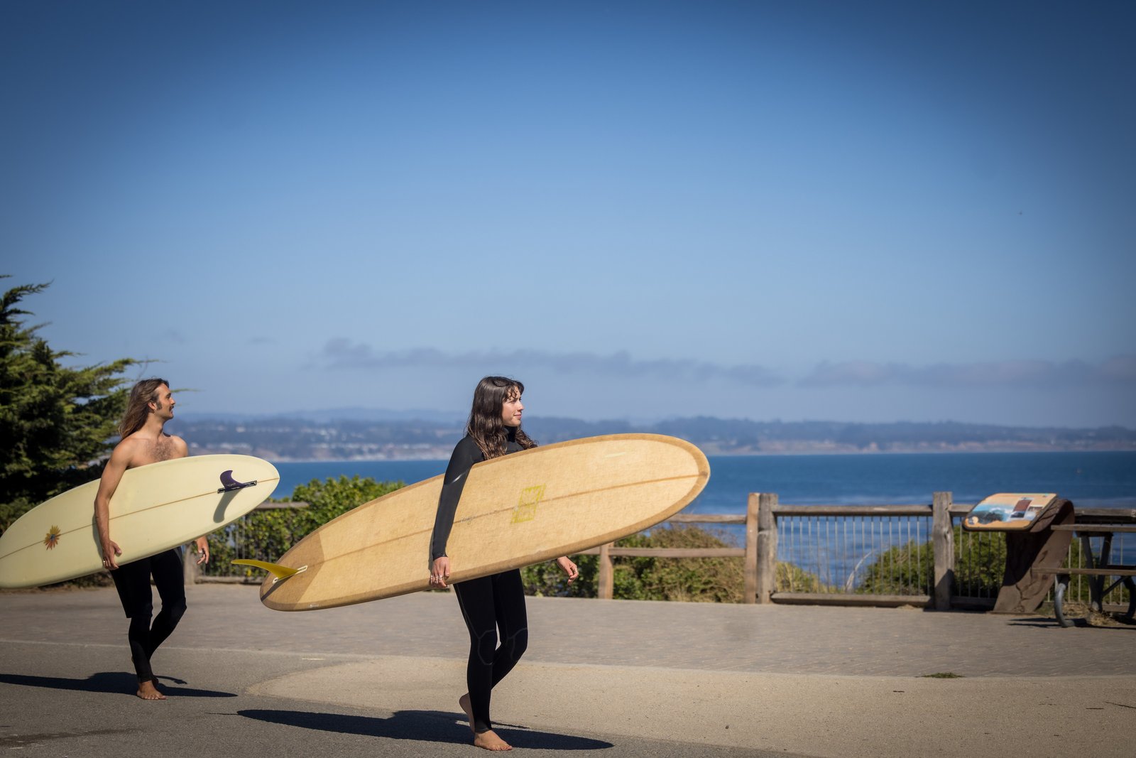 Two surfers carrying longboards along the coastal path beside Monterey Bay