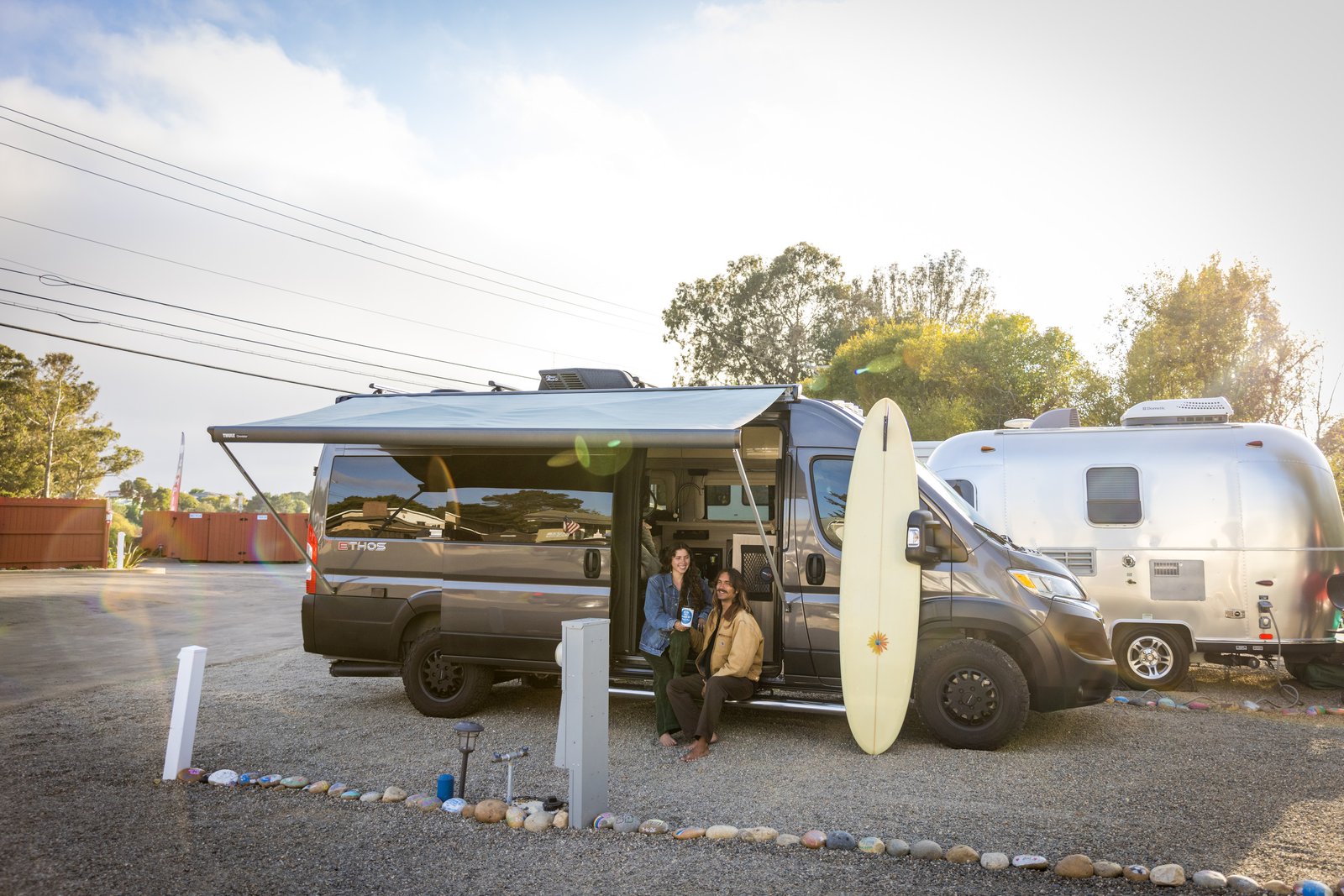 Two people sitting in a campervan doorway beneath an awning, with a longboard beside them