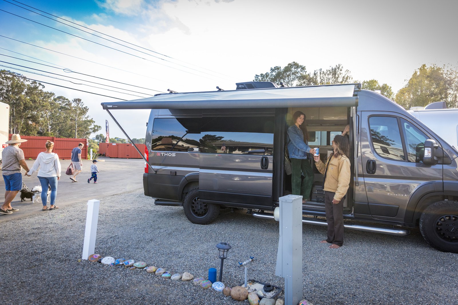 Two people at the open side door of a campervan, with other people and dogs walking through Beach RV Pleasure Point.