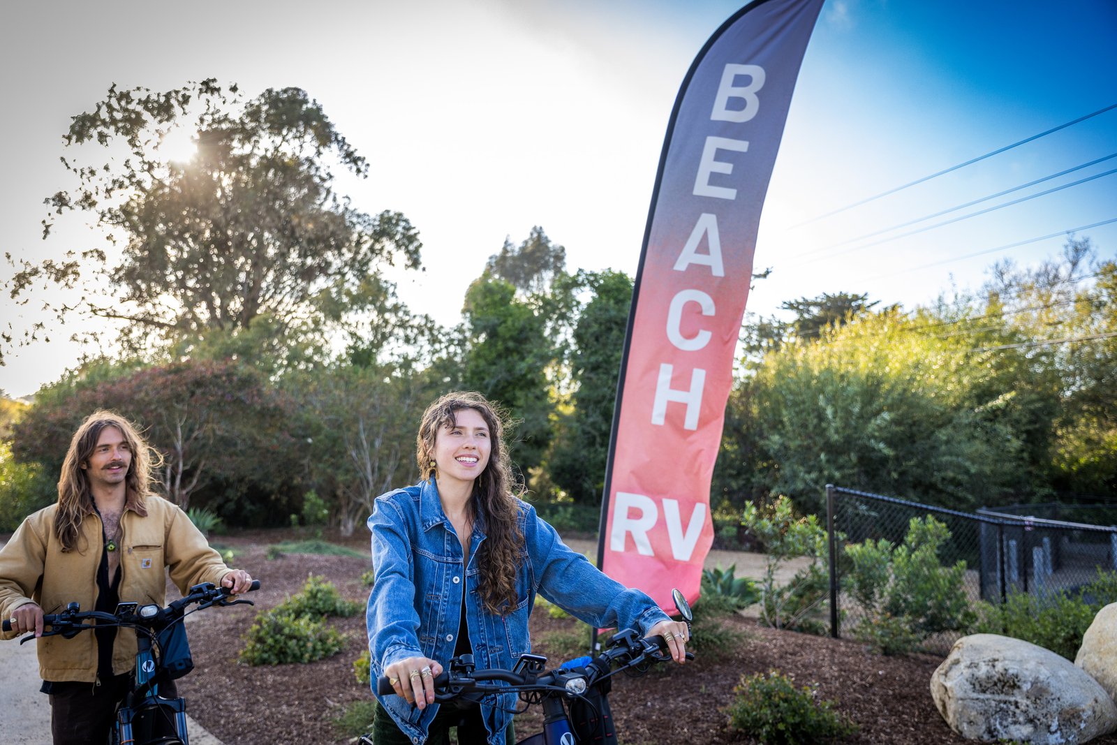 Two people on e-bikes beside the Beach RV entrance flag