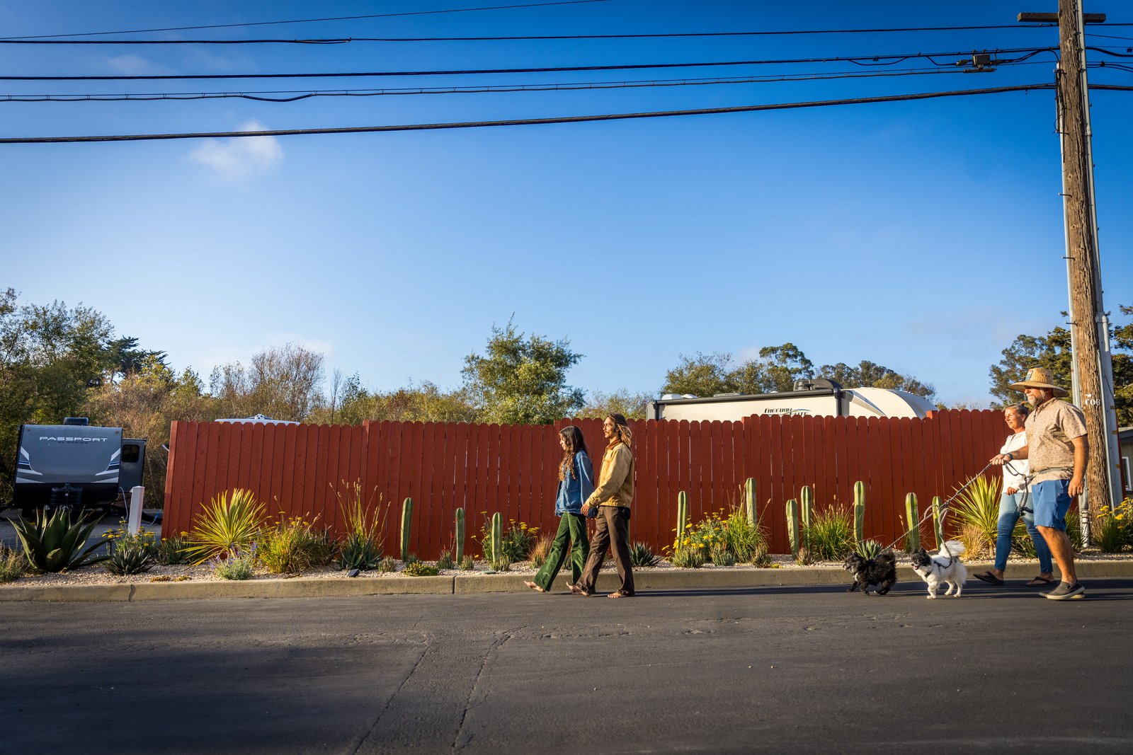 Four people and two dogs walk beside the red fence and garden at Beach RV Pleasure Point.