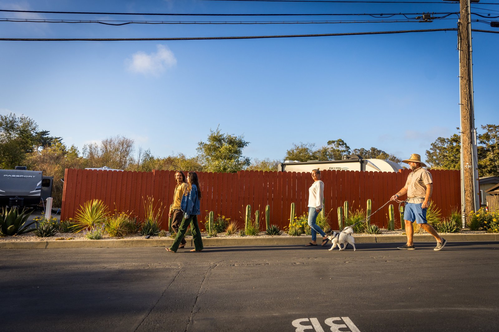 Four people walking dogs beside the red fence and garden at Beach RV Pleasure Point.
