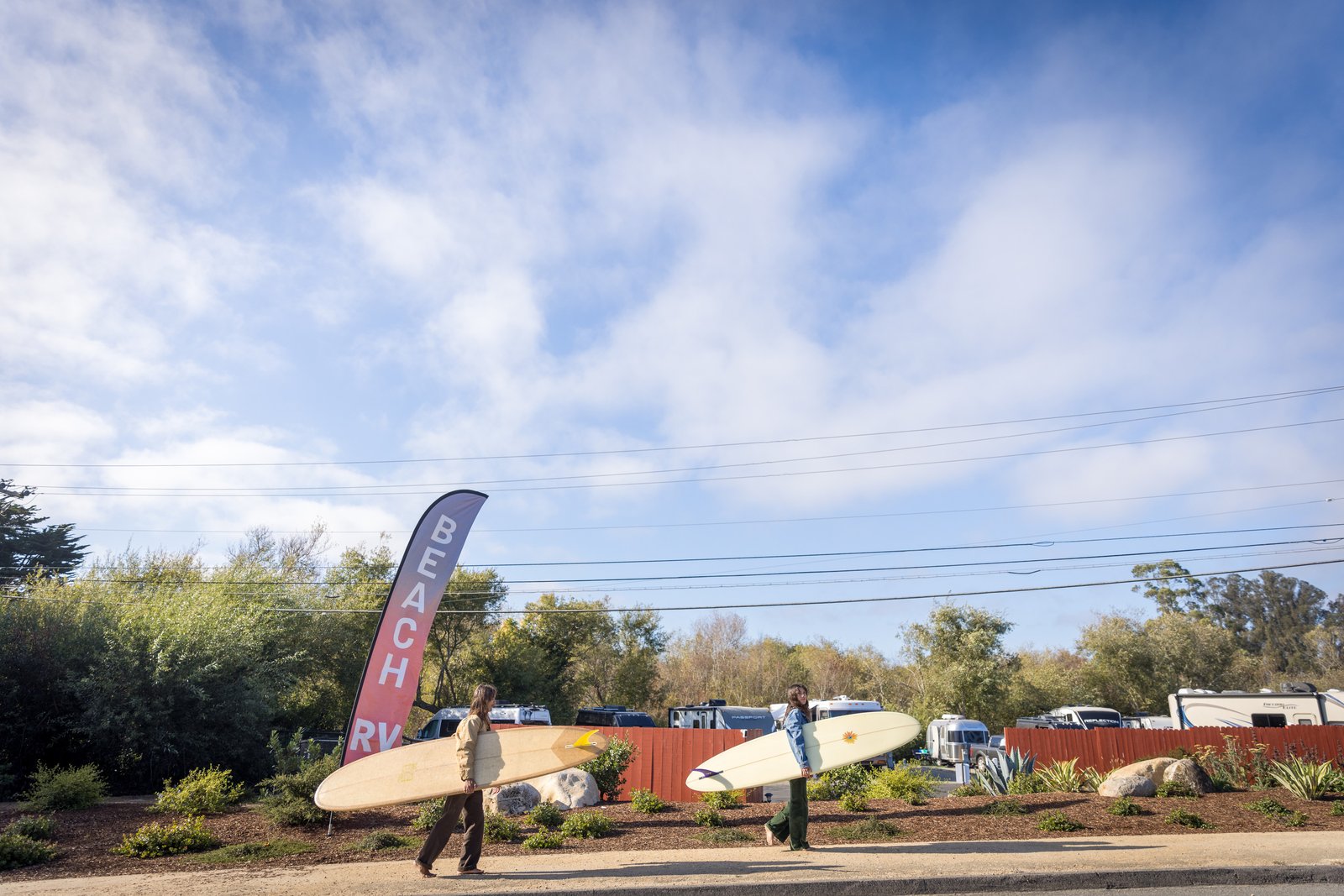 Two people carrying surfboards past a Beach RV flag and the park entrance.