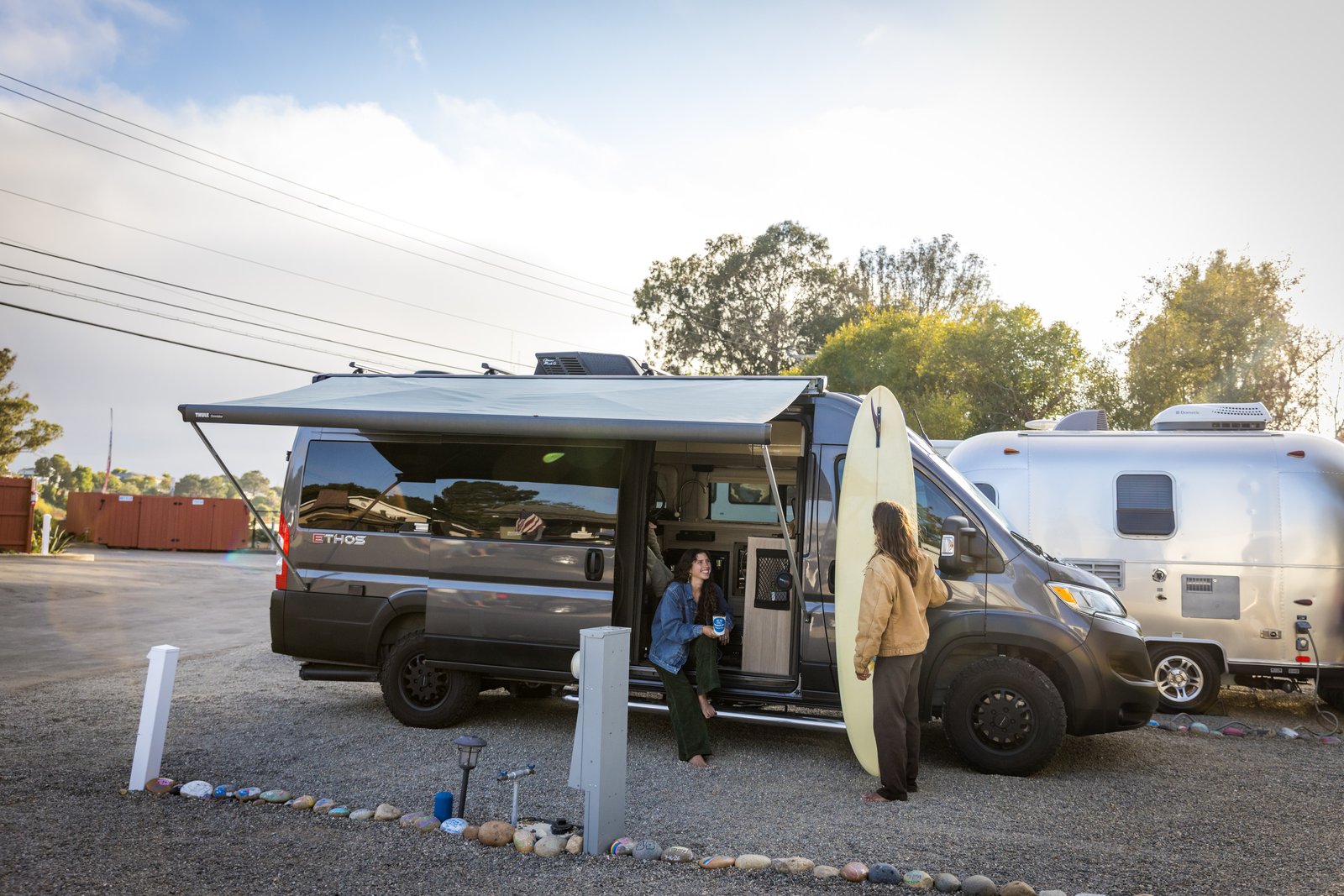 Two people beside an open campervan with its awning extended and surfboards nearby at Beach RV Pleasure Point.