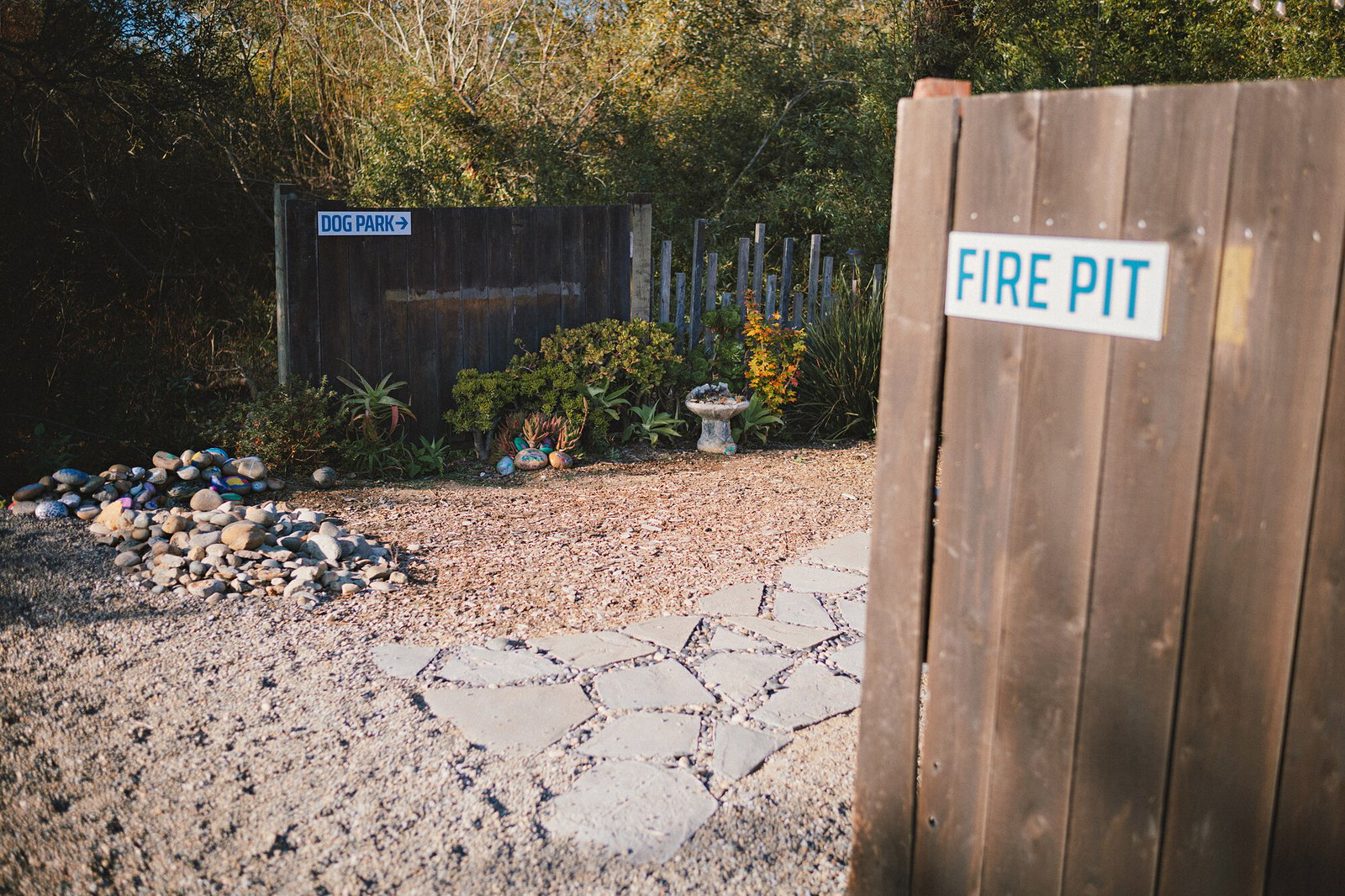 A “Fire Pit” sign beside a stone path, painted rocks and a sign pointing toward the dog park.