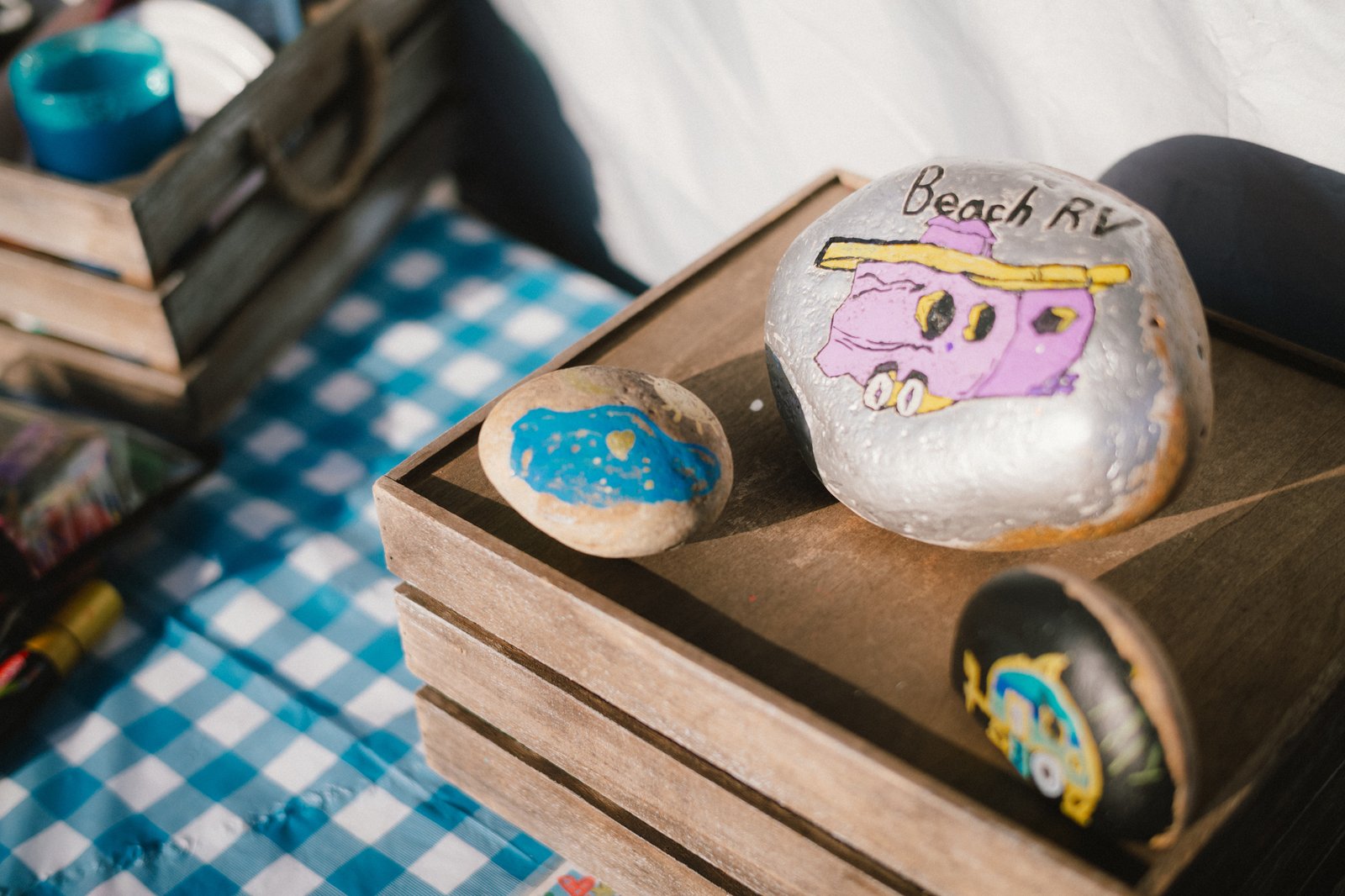 Guest-painted rocks displayed in wooden crates on a blue checked tablecloth at Beach RV Pleasure Point.