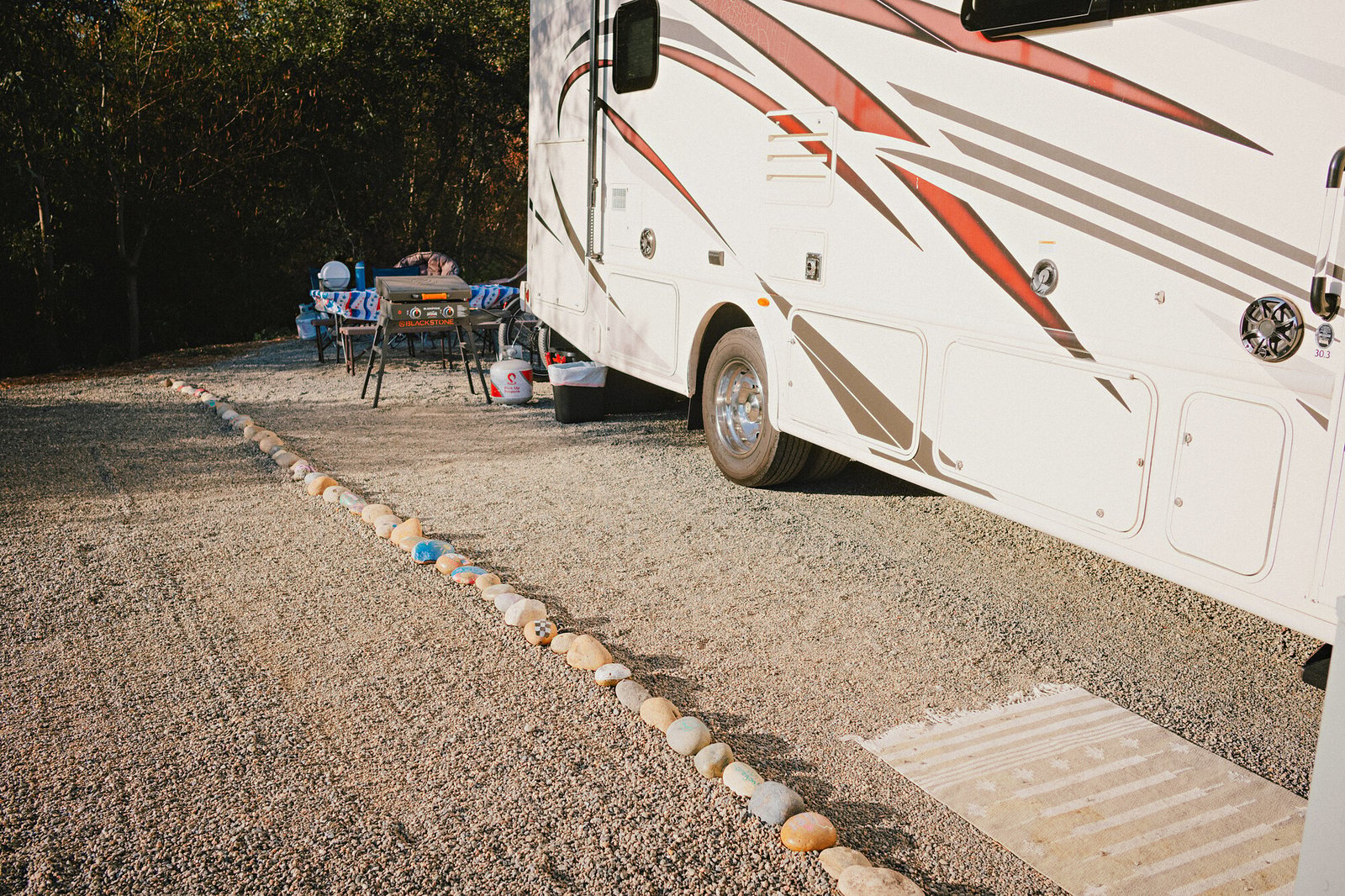 Class A motorhome and outdoor camp kitchen at golden hour beside the painted rock border