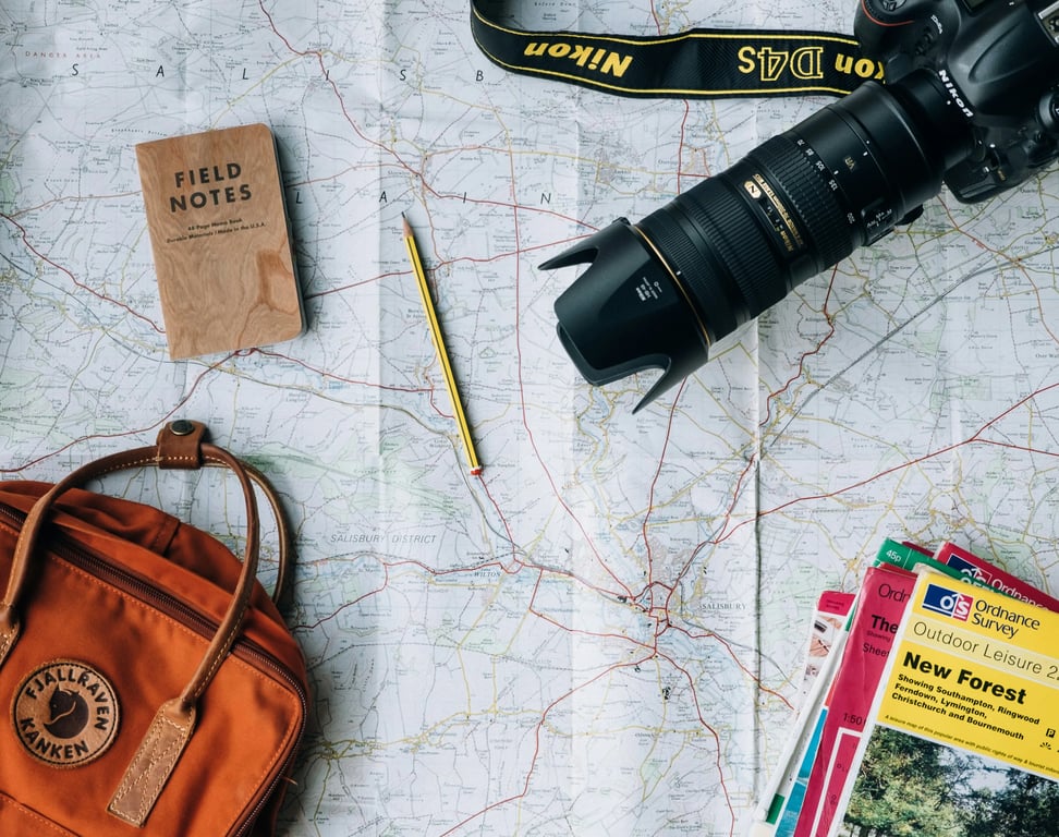Image of travel planning tools. A smartphone, camera, camera lens, watch, map, and ink pen on a dark wood table.