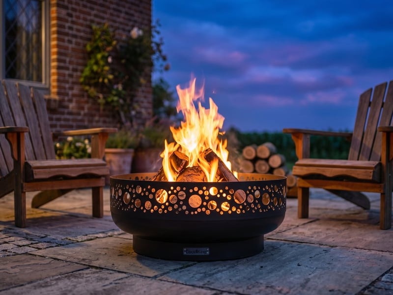 Decorative steel fire pit bowl lit with flames on a British brick-house patio at dusk with two empty chairs nearby