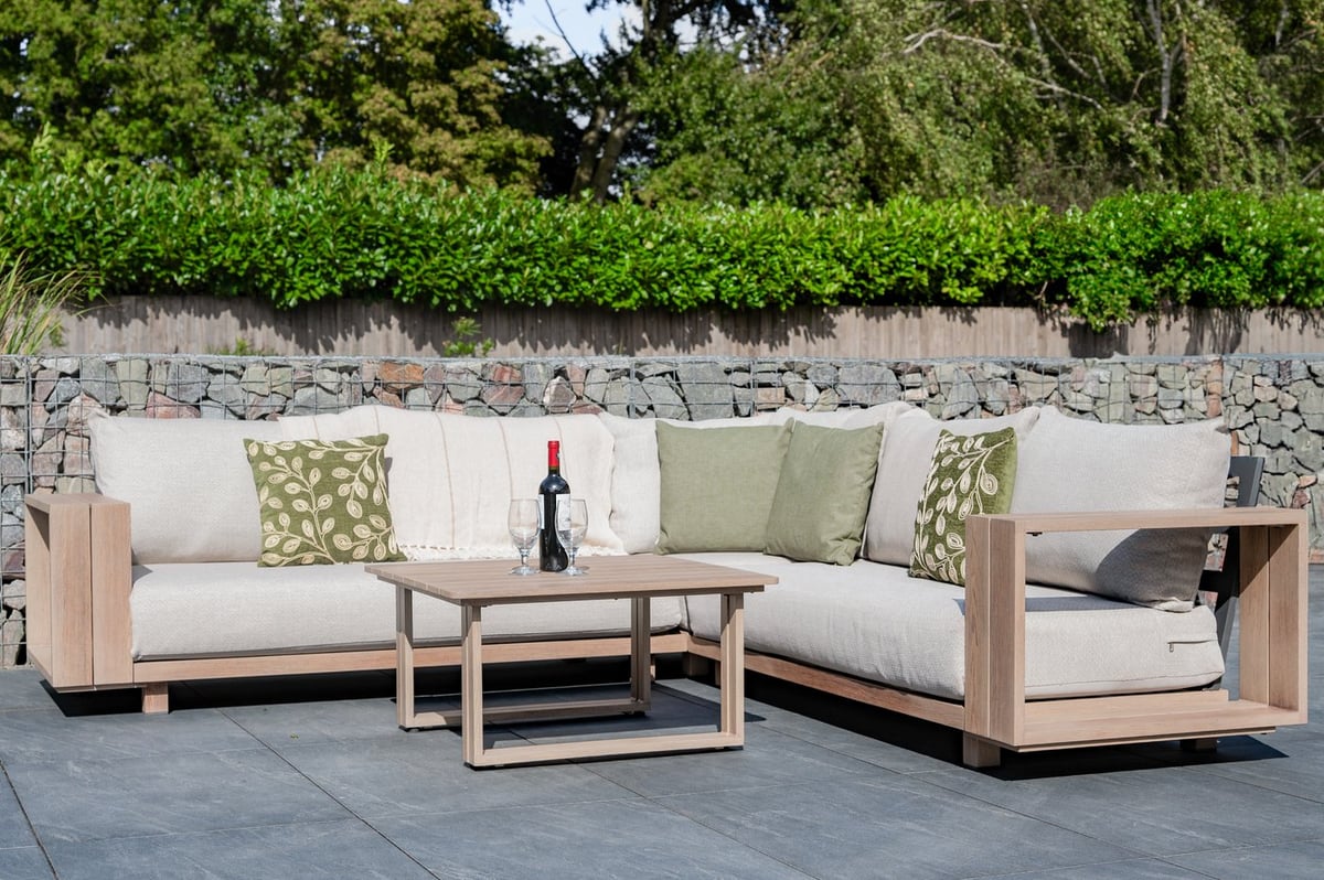 Cream-cushioned wood-frame garden corner sofa set on grey slate paving against a gabion stone wall and hedge in a narrow UK garden