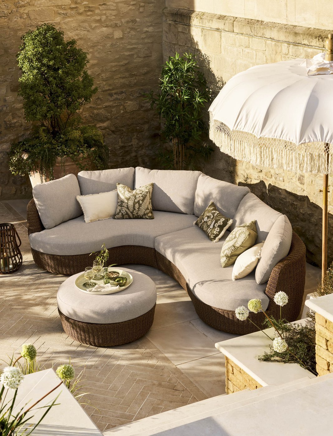 Curved garden corner sofa set in dark PE rattan with grey cushions and a fringed parasol in a honey-coloured stone walled courtyard