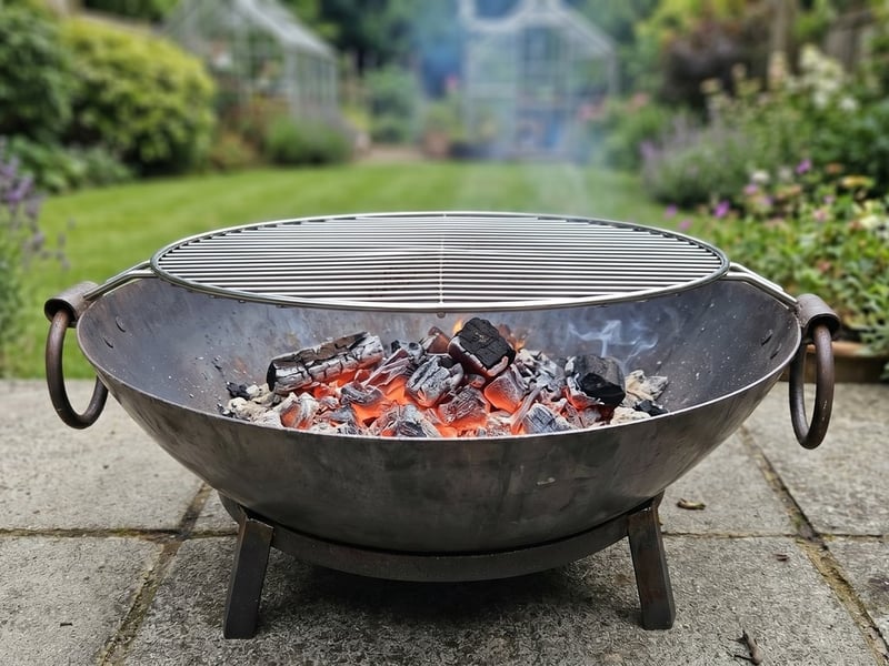 Steel Kadai fire pit bowl on a stand with a BBQ grill and glowing embers on a daytime UK patio