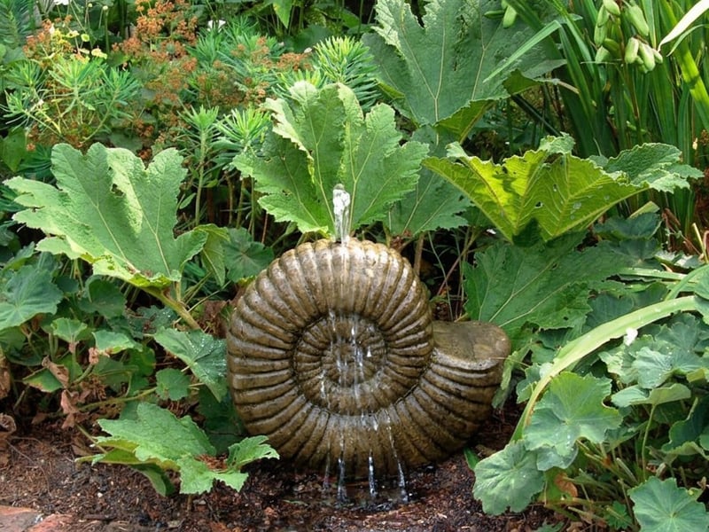 Grand Ammonite stone fountain in a UK shrub border with water cascading down the spiral fossil pattern