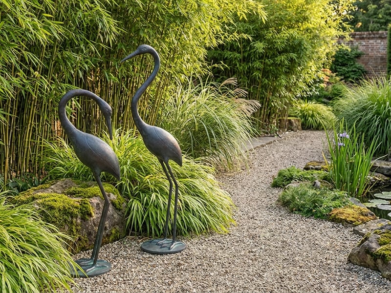 Pair of elegant bronze crane garden ornaments beside a gravel path in a UK garden with bamboo backdrop