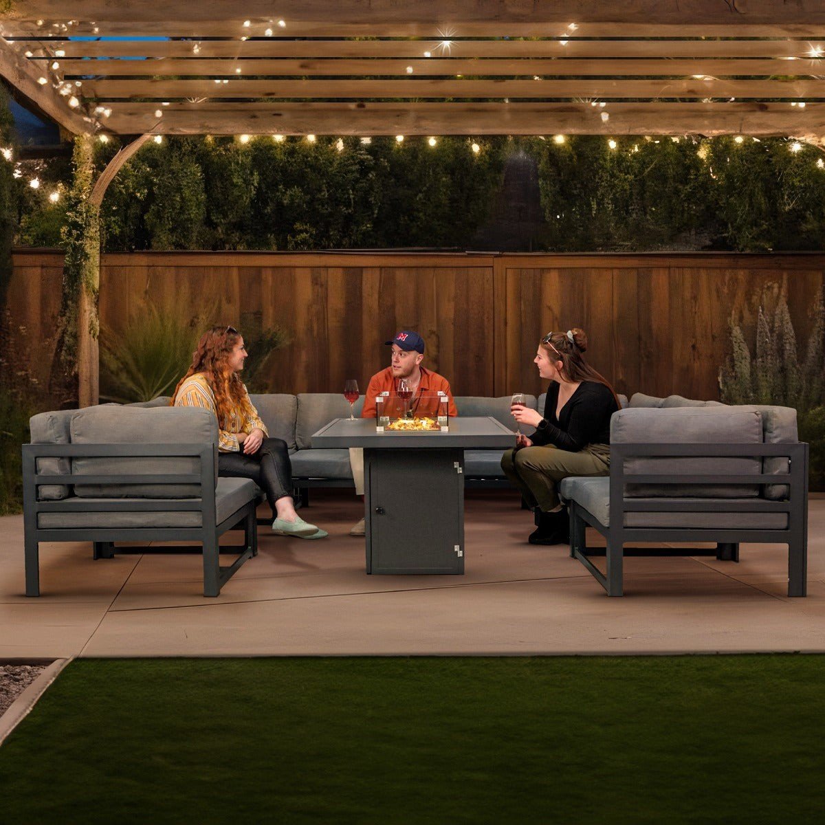 Anthracite aluminium modular garden corner sofa set arranged around a lit gas fire pit table under a pergola with string lights at dusk in a UK garden