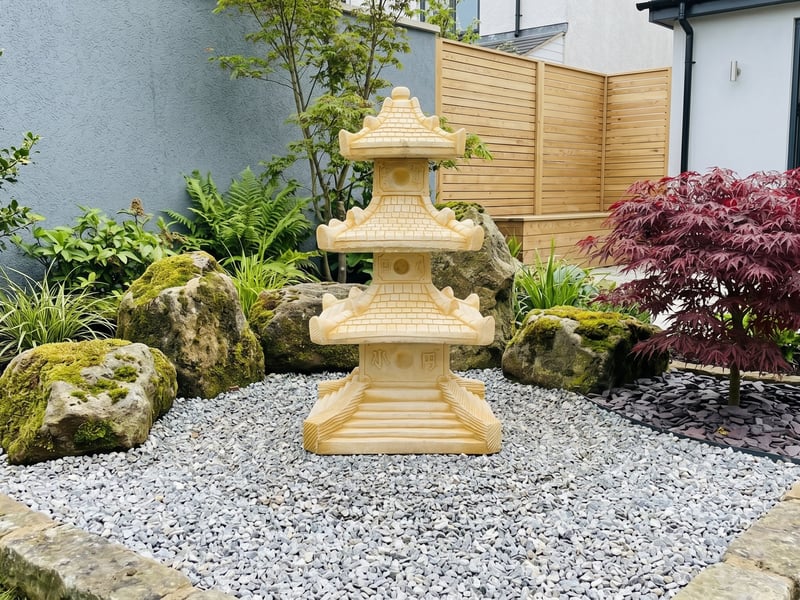 Three-tier sandstone pagoda garden ornament as a focal point on raked gravel in a modern UK courtyard garden with a red acer and mossy boulders