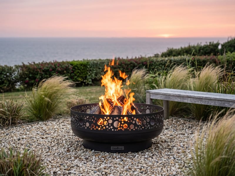 Cook King Boston 80cm decorative steel fire bowl lit at sunset in a Cornish coastal garden with ornamental grasses