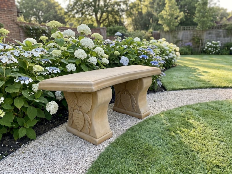 Curved Leaf stone garden bench in sandstone on a gravel path beside a hydrangea border in a suburban garden