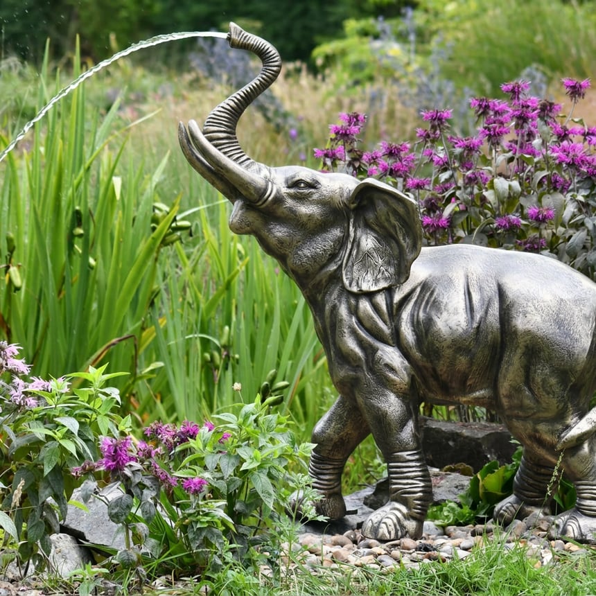 Baby elephant fountain in hand-finished pewter cast aluminium spraying water from its raised trunk beside garden planting