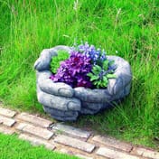 Cupped Hands Stone Garden Planter planted with purple and blue flowers in a garden setting