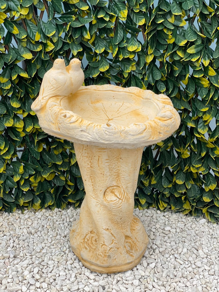 Dove bird bath in sand stone with two sculpted doves on the bowl rim and a tree-trunk pedestal in a UK garden