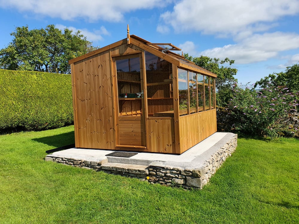 Swallow Rook 8ft 9in wide ThermoWood potting shed greenhouse on a stone-faced base in a garden, with a solid gable end and glazed working side