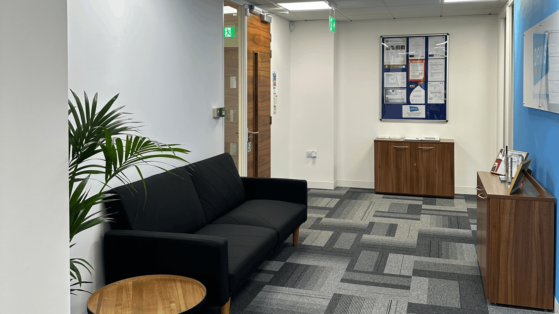 Reception and waiting area for the license check office fit out with black sofa, plant, timber storage units, blue feature wall, acrylic signage and grey patterned carpet tiles