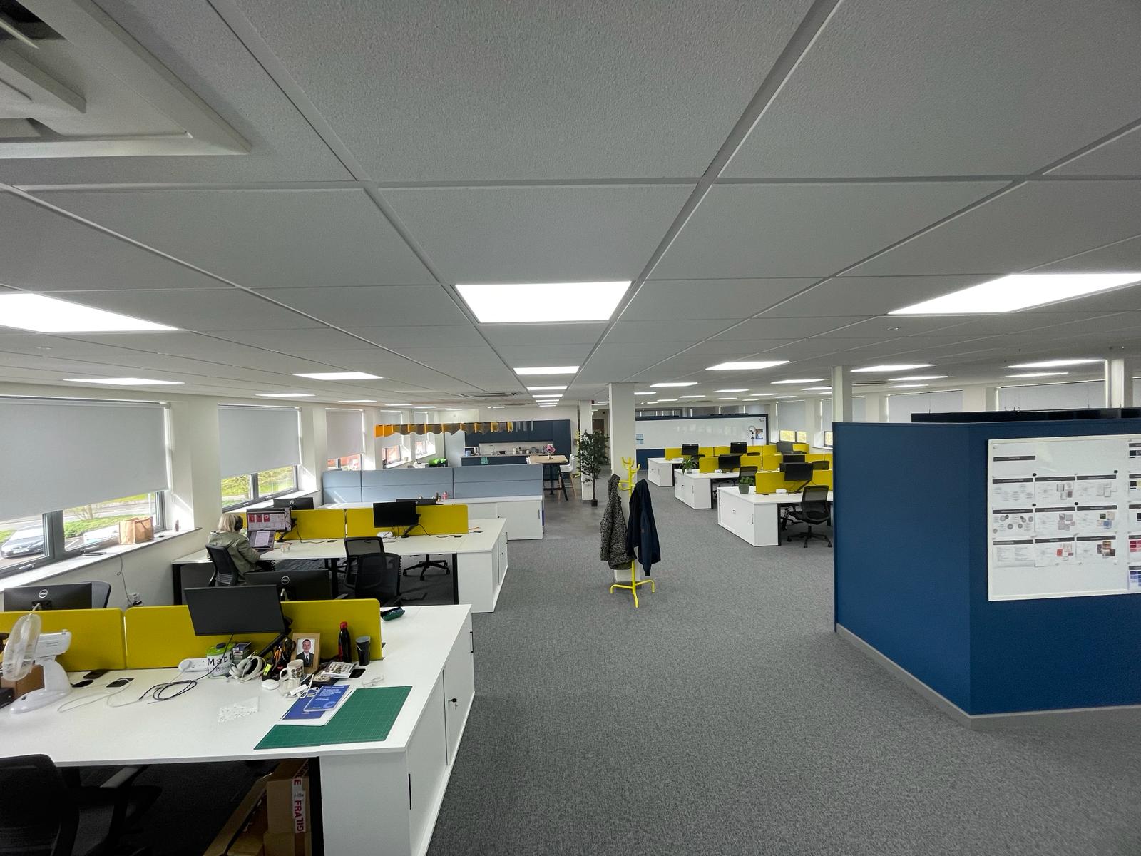 Open plan office at card factory wakefield showing white bench desks, yellow acoustic screens, blue feature walls and grey carpet tiles