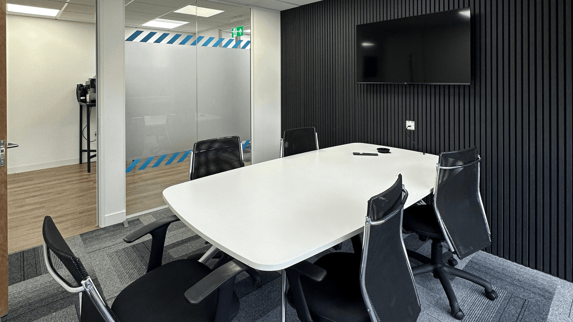 Completed license check meeting room with white meeting table, black mesh chairs, black acoustic slat wall, wall-mounted tv and glazed partitioning to the breakout area