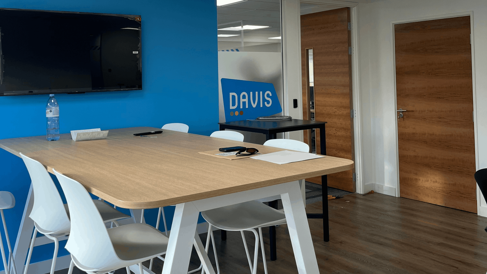License check office fit out breakout space showing high meeting table, white stools, blue branded feature wall, wall-mounted tv, timber doors and wood-effect flooring