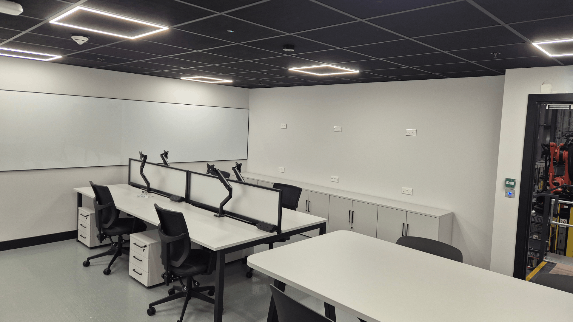 Open plan office area inside cimcorp mezzanine with white desks, black operator chairs, monitor arms, storage units, rubber flooring and black suspended ceiling