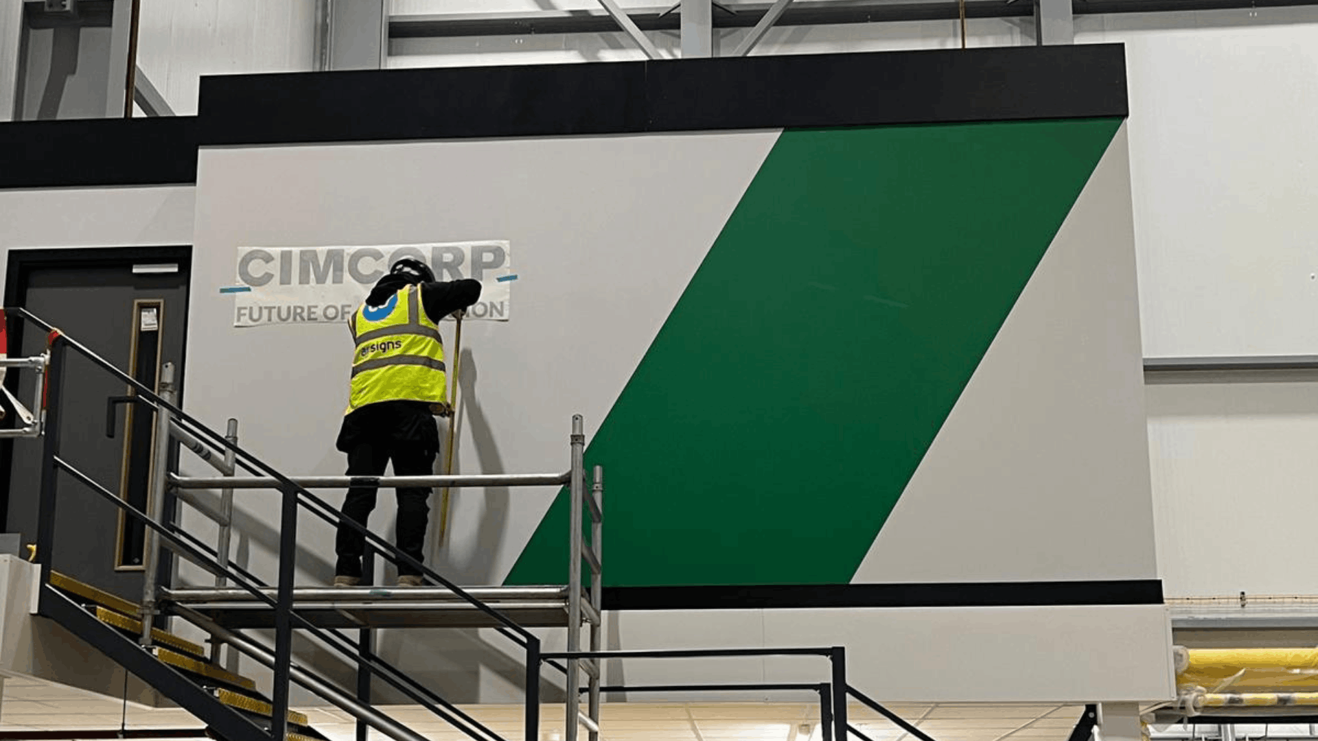 Installer applying cimcorp wall branding to the exterior of the mezzanine office beside the black staircase during the fit out process
