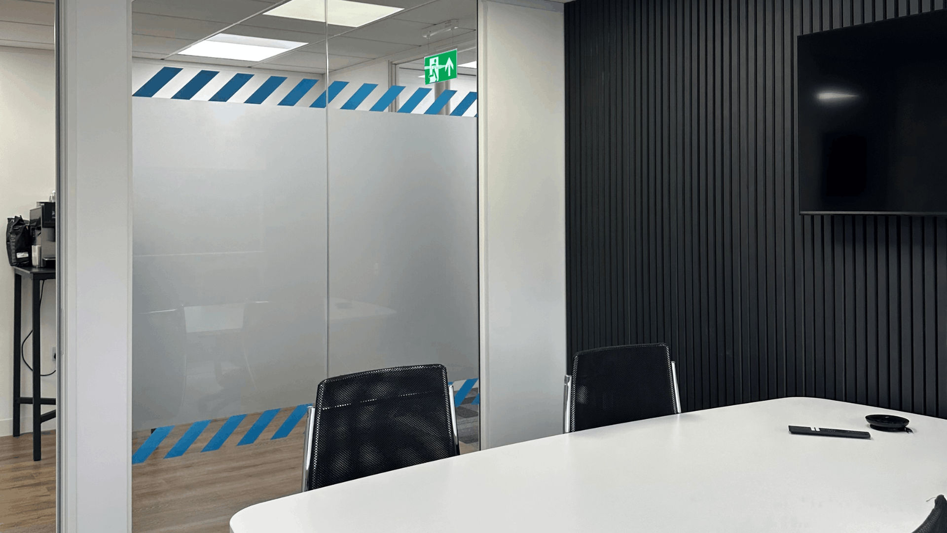 Completed license check meeting room with black acoustic slat wall, wall-mounted tv, white meeting table, black chairs and glazed partitioning with blue manifestation