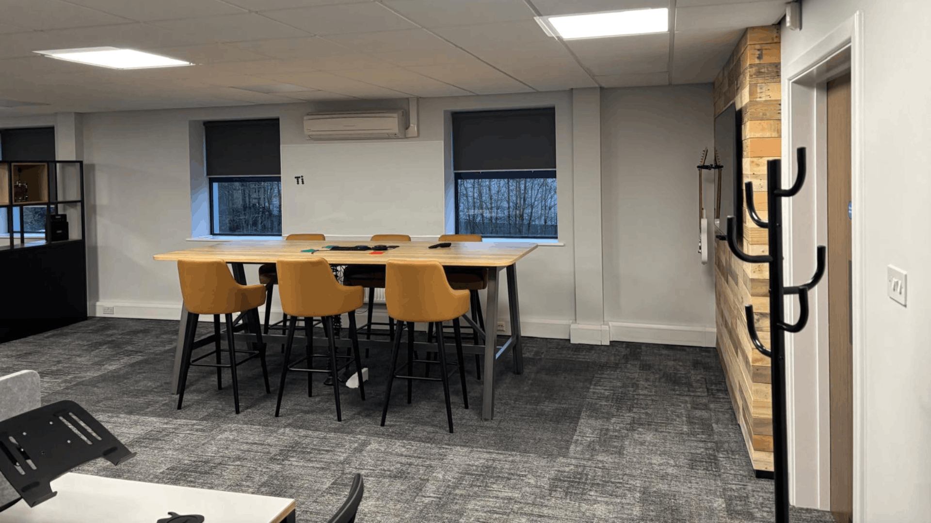 Refurbished open-plan office meeting area with poseur-height table, tan chairs, grey carpet tiles and timber feature wall