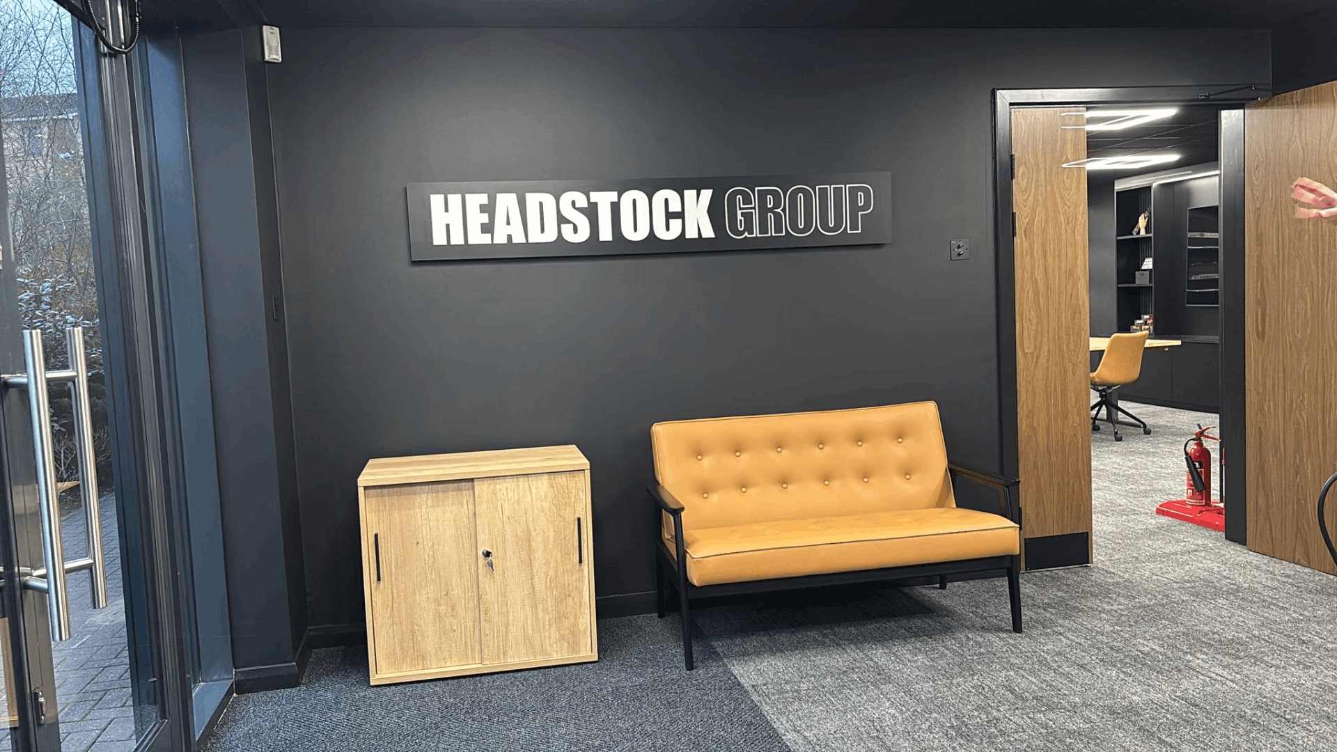 Headstock group reception area with illuminated wall signage, tan sofa, timber storage unit and dark feature wall