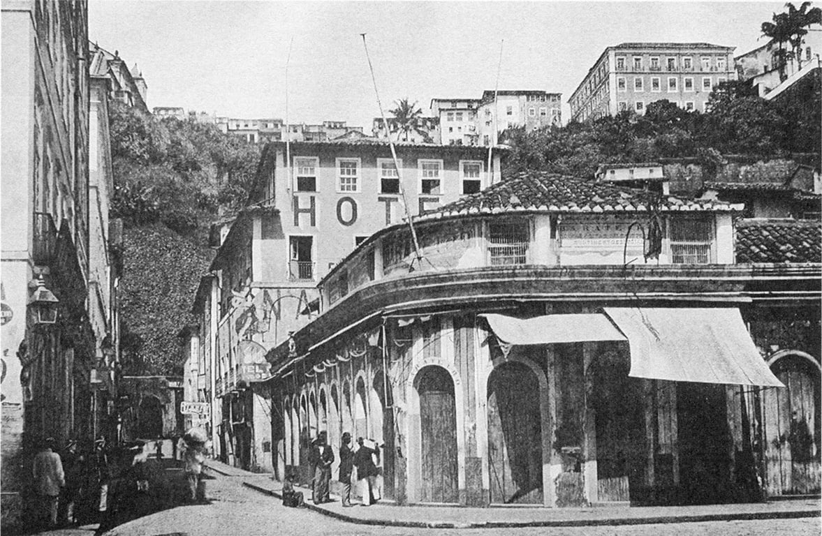 Fotografia histórica dos Arcos de Santa Bárbara, de 1885, localizada na antiga Rua Santa Bárbara, no bairro do Comércio, em Salvador. A imagem mostra uma esquina formada por um prédio térreo de arquitetura colonial, com uma sequência de arcos voltados para a rua e cobertura de telhas cerâmicas. Sobre o edifício, uma construção de vários andares exibe, em letras grandes na fachada, a palavra “HOTEL”. Na calçada, algumas pessoas estão reunidas próximas aos arcos, enquanto outras circulam pela rua. À esquerda, aparecem mais edifícios alinhados ao longo da via. Ao fundo, o terreno sobe em direção à parte alta da cidade, com diversos casarões e prédios distribuídos pela encosta. A fotografia registra a região do antigo Mercado de Santa Bárbara, um dos principais espaços de circulação de mercadorias e de atuação dos ganhadores na Salvador do século XIX.