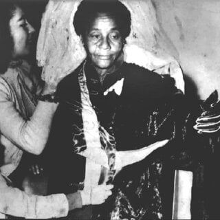 A imagem mostra uma de Maria José Bezerra, conhecida como Maria Soldado, de pele negra, em primeiro plano, sendo auxiliada por outra pessoa que aparece parcialmente à esquerda. Foi uma combatente negra da Revolução Constitucionalista de 1932 e está vestida com um tecido escuro que parece ser colocado ou ajustado sobre seus ombros. Seu semblante é sério e sereno, com o olhar levemente direcionado para baixo. A pessoa ao lado, cuja face aparece apenas parcialmente, parece estar acomodando ou organizando a vestimenta. O fundo é simples, sem detalhes específicos, reforçando o foco nas duas figuras, seja no gesto de cuidado ou preparação.