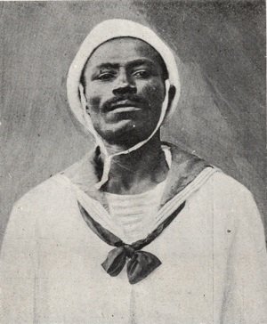 Fotografia em preto e branco de João Cândido Felisberto, líder da Revolta da Chibata, vestindo uniforme de marinheiro. Ele está de frente, com expressão firme e olhar direto. Usa um gorro branco preso sob o queixo por uma fita e uma blusa de gola larga com lenço escuro amarrado na frente, típico do uniforme naval da época. O fundo é neutro, escuro e sem elementos visuais, destacando a figura de João Cândido. A imagem reforça sua identidade como marinheiro e símbolo de resistência contra os castigos físicos e o racismo institucionalizado na Marinha brasileira.