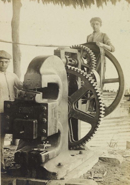Fotografia em sépia capturando dois meninos sobre máquina de cortar ferro e aço, Cruzeiro do Sul, Acre. A composição da cena apresenta uma máquina industrial robusta, ocupando a maior parte do enquadramento, com dois jovens posicionados em lados opostos. O menino à esquerda está encostado na máquina e observa a câmera, enquanto o da direita segura o volante grande da máquina. A ação é estática, capturando um momento de pausa, provavelmente para a foto. A estrutura da cena sugere um ambiente de trabalho, possivelmente uma fábrica ou oficina.