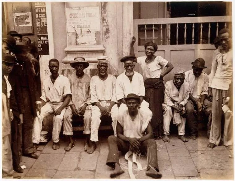 Fotografia histórica de ganhadores de Salvador, pertencente ao acervo da The New York Public Library. A imagem mostra um grupo de homens e uma mulher negros reunidos em frente à fachada de um edifício. No centro, seis homens estão sentados em um banco, enquanto outro permanece sentado no chão, com os braços apoiados sobre as pernas. Ao lado deles, uma mulher está em pé, com uma das mãos apoiada na cintura. Outros homens aparecem de pé nas laterais da fotografia, alguns usando chapéus e observando a câmera. A maioria veste roupas claras e simples, como camisas de mangas curtas, calças e chapéus ou boinas. Ao fundo, a fachada apresenta portas, janelas e cartazes afixados na parede. A fotografia registra trabalhadores conhecidos como ganhadores, responsáveis pelo transporte de mercadorias, bagagens e outros serviços nas ruas de Salvador durante o século XIX.