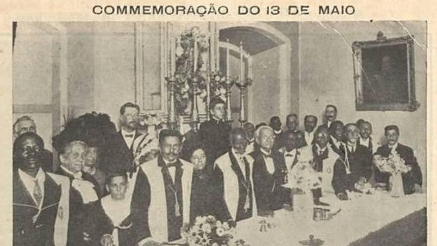 Fotografia em preto e branco de uma cerimônia na Igreja Nossa Senhora do Rosário, no Rio de Janeiro, em 1911, com o título 'Comemoração do 13 de Maio' impresso na parte superior. Ao longo de uma mesa comprida, diversas pessoas negras e brancas estão sentadas ou em pé. Entre elas, homens usando fraques e gravatas, alguns com colares e medalhas, e mulheres com vestidos escuros. No centro, está o ex-presidente do conselho de ministros João Alfredo e Maria Henriqueta Sena, a viúva do abolicionista José do Patrocínio. A mesa está decorada com vasos de flores e utensílios de cerâmica. Ao fundo, nota-se um altar ricamente adornado e, à direita, um quadro pendurado na parede. A imagem mostra a participação ativa da população negra em cerimônias e organizações sociais do período pós-abolição.