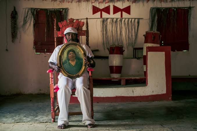 Fotografia de um filho de santo do Terreiro Ilê Axé Icimimó Aganju Didè, de Vinicius Xavier, publicada pela FotoDoc. A imagem mostra um homem negro sentado em uma cadeira de madeira, no centro de um salão de terreiro. Ele veste roupas totalmente brancas, usa um gorro branco na cabeça e está descalço. Com as duas mãos, segura um quadro oval com a fotografia de uma mulher. Ao fundo, aparecem atabaques, bancos, elementos decorativos e símbolos religiosos nas cores vermelha e branca, além de janelas com venezianas de madeira. A fotografia registra o interior do terreiro e destaca a presença e a continuidade das tradições religiosas de matriz africana.
