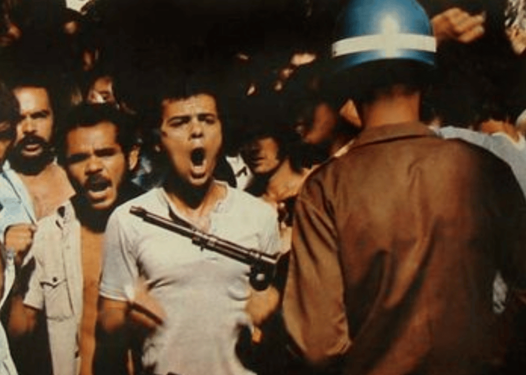 Fotografia colorida mostra um operário em ato de resistência enfrentando um policial militar durante uma manifestação. O trabalhador, de camisa branca, está à frente de uma multidão e grita com expressão de coragem, mesmo com o cano de um fuzil apontado diretamente para seu peito. O policial, de costas, usa capacete azul e uniforme marrom. Ao fundo, outros manifestantes acompanham a cena com rostos tensos e indignados. A imagem foi registrada em maio de 1980, em São Bernardo do Campo (SP), durante as greves dos metalúrgicos contra a ditadura.