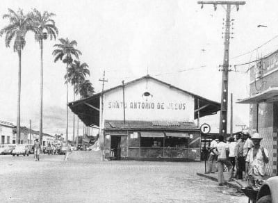 Fotografia em preto e branco da estação ferroviária de Santo Antônio de Jesus, na Bahia, registrada no século XX. A imagem mostra o prédio principal da estação ao centro, com cobertura ampla e a inscrição “Santo Antônio de Jesus” na fachada. Em frente ao edifício, há uma área aberta e pavimentada. À esquerda, uma fileira de altas palmeiras acompanha a avenida, onde aparecem alguns automóveis estacionados e pessoas caminhando. À direita, um grupo de pessoas permanece na calçada próximo a um poste de energia e a outros edifícios comerciais. A fotografia registra um período em que a estação ferroviária era um importante ponto de circulação de passageiros e mercadorias no município.