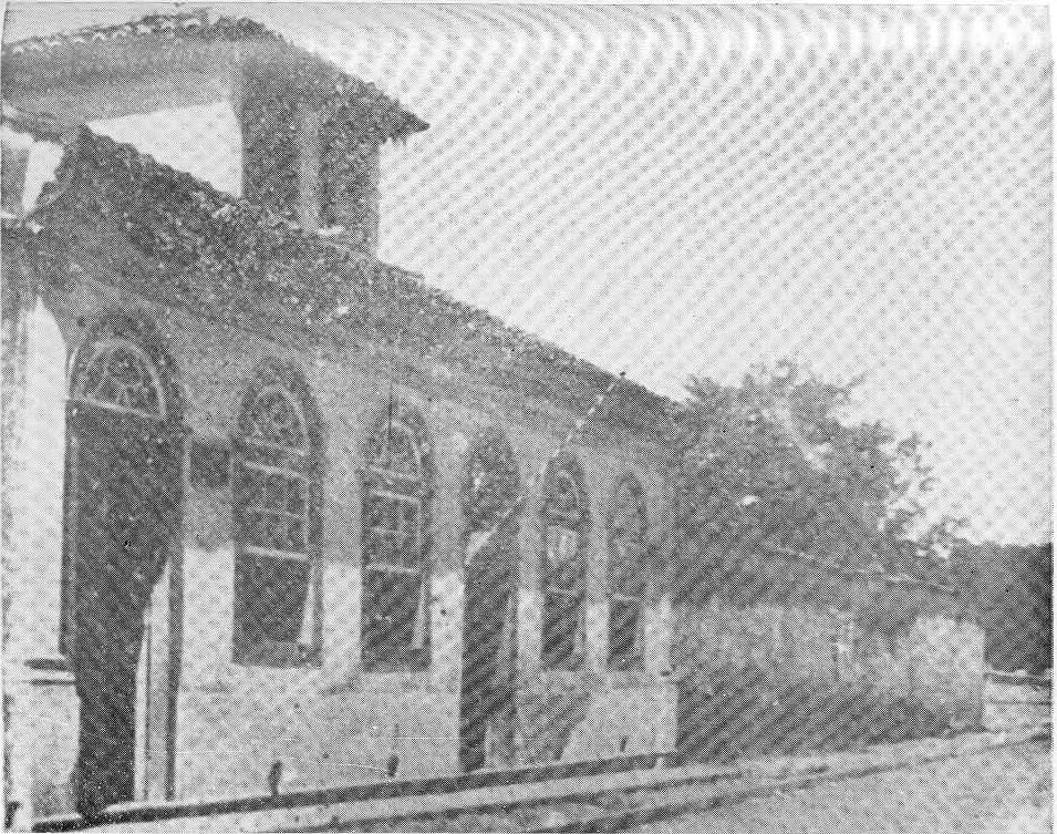 Fotografia em preto e branco de um antigo prédio histórico com fachada colonial. À esquerda, vê-se uma construção de um andar, com quatro grandes janelas em arco e uma porta com moldura arredondada. As janelas têm grades e caixilhos de vidro. O telhado é de telhas cerâmicas, e uma pequena torre ou campanário se eleva acima da construção principal. À direita da imagem, há um muro baixo e vegetação ao fundo. A calçada acompanha o contorno da construção. A imagem tem aspecto granulado, típico de impressões antigas. Trata-se do antigo prédio do Colégio São Benedito, situado na Avenida Moraes Salles.