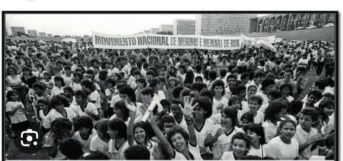 A fotografia colorida mostra várias crianças e adolescentes caminhando em fila durante uma manifestação em Brasília. Eles seguram faixas e cartazes. A composição é frontal, capturando o movimento coletivo em ambiente externo urbano. As ações são de manifestar, protestar e reivindicar direitos.