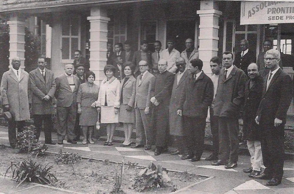 Fotografia em preto e branco feita em 1956, durante a inauguração da sede do clube social negro Satélite Prontidão, em Porto Alegre. Um grupo de homens e mulheres posa enfileirado em frente ao prédio, com colunas brancas e varanda, trajando roupas formais da época – ternos, sobretudos, gravatas e vestidos sociais. Entre eles estão figuras importantes: o compositor Lupicínio Rodrigues (penúltimo à direita), além dos políticos Carlos Santos e Alceu Collares (posicionados ao lado de um padre, no centro da imagem). Ao fundo, sob a varanda, outras pessoas observam a cena. Na fachada, vê-se parcialmente a placa com os dizeres Associação Satélite Prontidão – Sede Própria.
