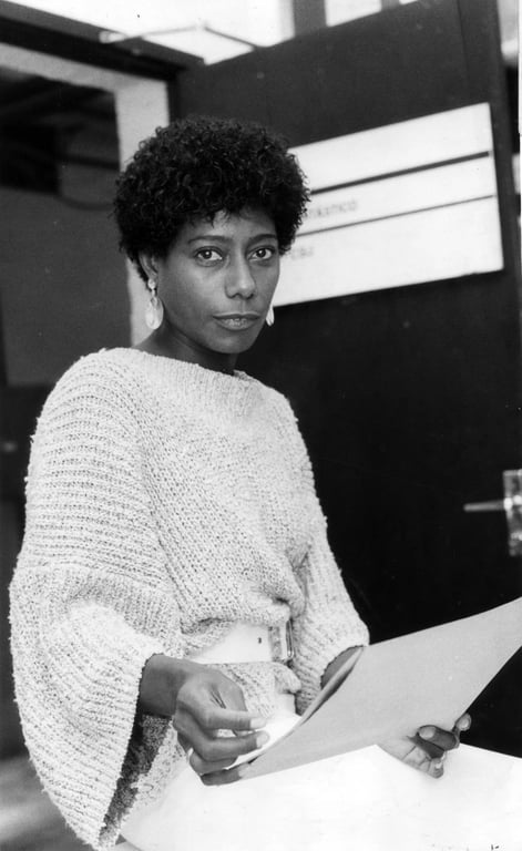 A fotografia em preto e branco retrata a repórter Glória Maria, em pé perto de uma porta, segurando um documento e olhando para a câmera. Ela está centralizada no enquadramento, com o corpo ligeiramente inclinado para a direita, criando uma impressão de serenidade e firmeza. Atrás dela, há uma porta escura, com um letreiro branco com texto, e uma abertura à esquerda, sugerindo um espaço interior. A composição é simples e direta, focando nela e em sua expressão, e no documento que ela segura. A luz parece natural, vindo de uma fonte fora do quadro, projetando sombras sutis e definindo a textura dos materiais. Ela tem pele escura, e cabelos crespos e curtos. Usa brincos pendurados e um suéter de malha claro com mangas largas. Ela está vestida com uma saia branca e um cinto. Seus olhos são expressivos e seu olhar é direto para a câmera, transmitindo uma sensação de confiança e inteligência.