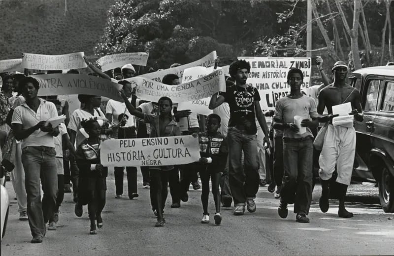 A fotografia em preto e branco captura um protesto político, com um grupo de pessoas NEGRAS marchando em uma rua, carregando cartazes e panfletos. Os sujeitos principais são os manifestantes, homens, mulheres e crianças NEGRAS de diferentes idades e vestimentas. Alguns usam roupas casuais, como camisas e calças jeans, enquanto outros parecem mais formais. As expressões faciais são variadas, algumas sérias e concentradas, outras transmitindo determinação e indignação. Os cartazes exibem mensagens de protesto e reivindicações.